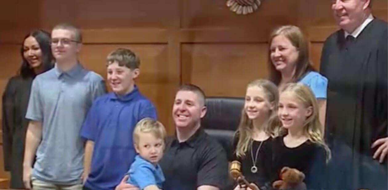 Group photo in a courtroom showing an adult couple with five children of various ages, standing with a judge in a black robe.