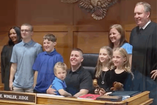 Group photo in a courtroom showing an adult couple with five children of various ages, standing with a judge in a black robe.