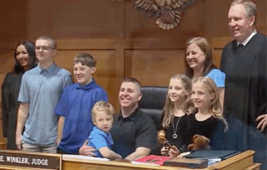 Group photo in a courtroom showing an adult couple with five children of various ages, standing with a judge in a black robe.