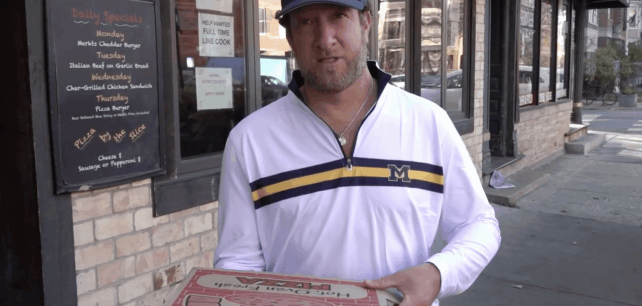 Dave Portnoy standing outside a brick storefront holding a pizza box during a pizza review.