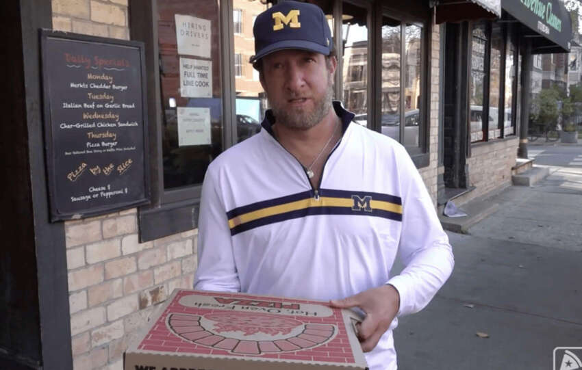 Dave Portnoy standing outside a brick storefront holding a pizza box during a pizza review.