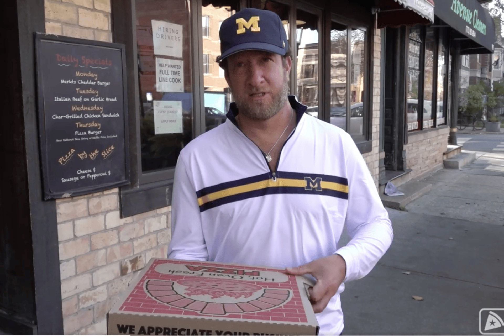 Dave Portnoy standing outside a brick storefront holding a pizza box during a pizza review.