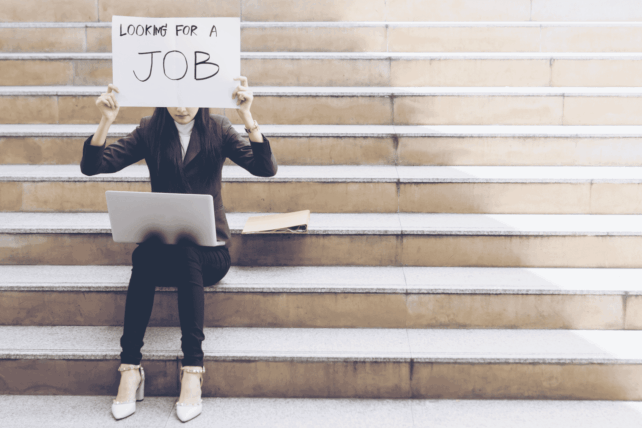 Job seeker sitting on steps holding a sign, illustrating the rising US unemployment rate in November 2025.
