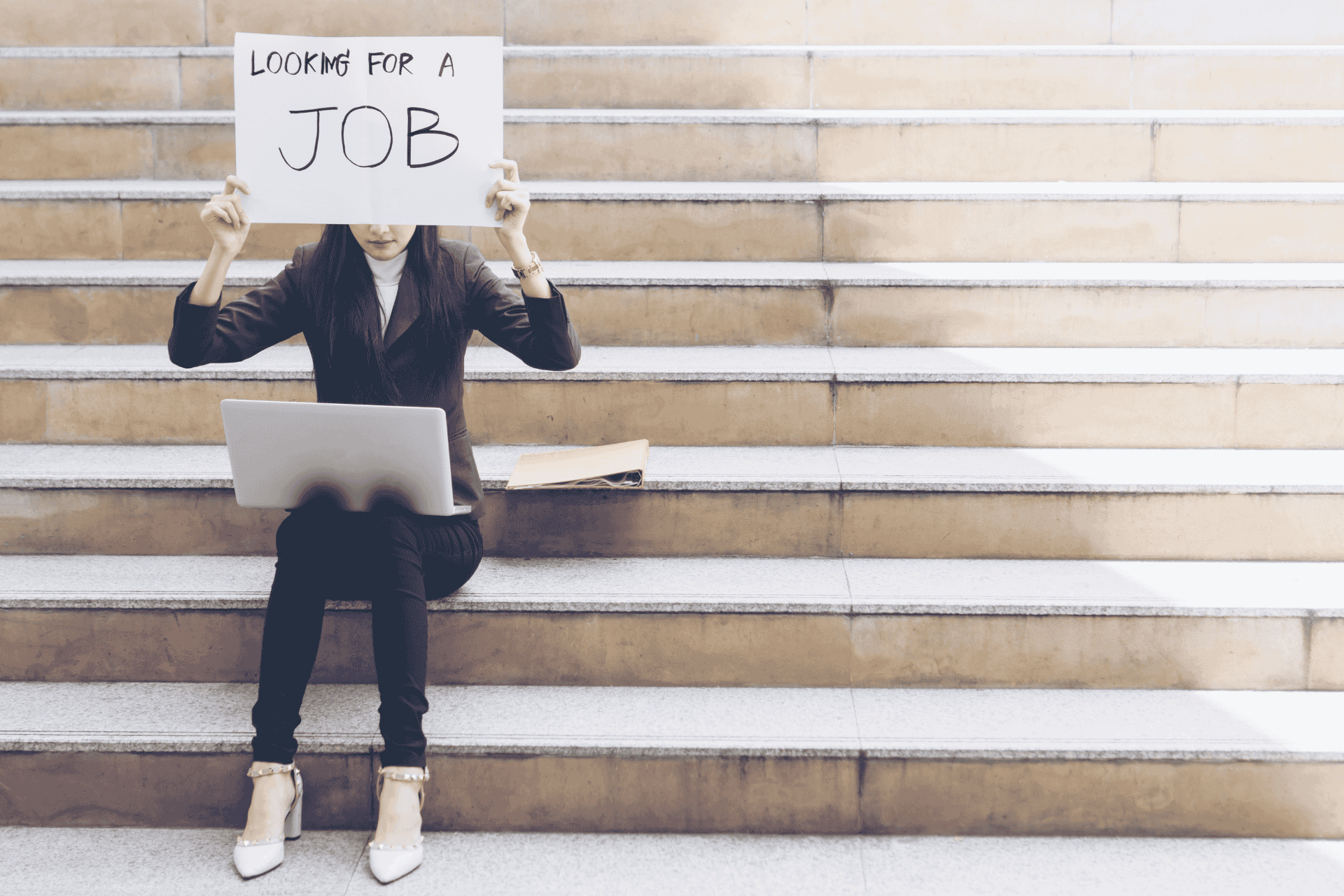 Job seeker sitting on steps holding a sign, illustrating the rising US unemployment rate in November 2025.
