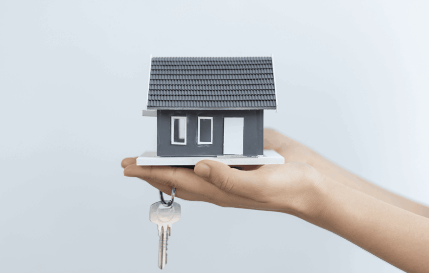 A person's hand holding a small model house with keys dangling from it, symbolizing home ownership or housing funding.