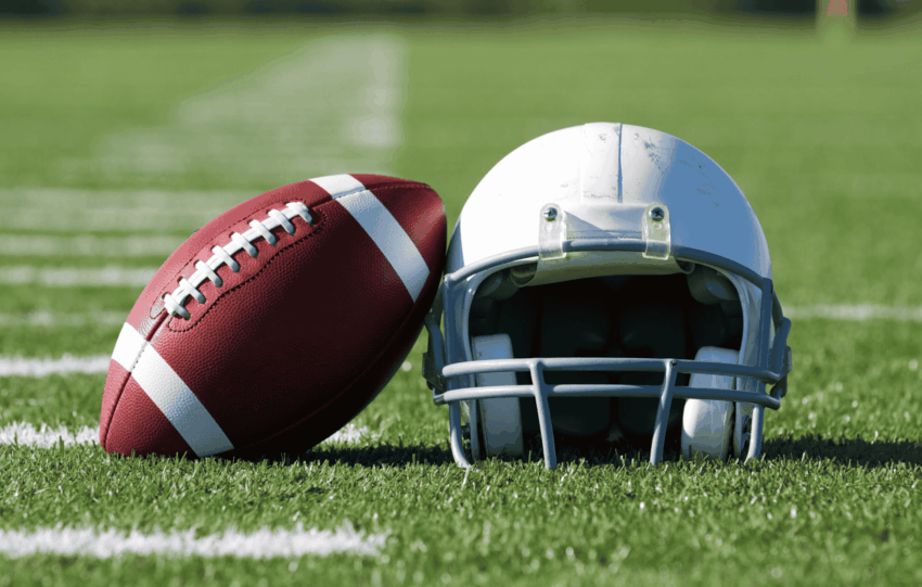 A close-up of an American football resting against a white football helmet on the grass of a football field.