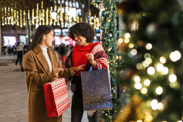 Two women holding shopping bags and smiling while standing outdoors at night surrounded by festive holiday lights.