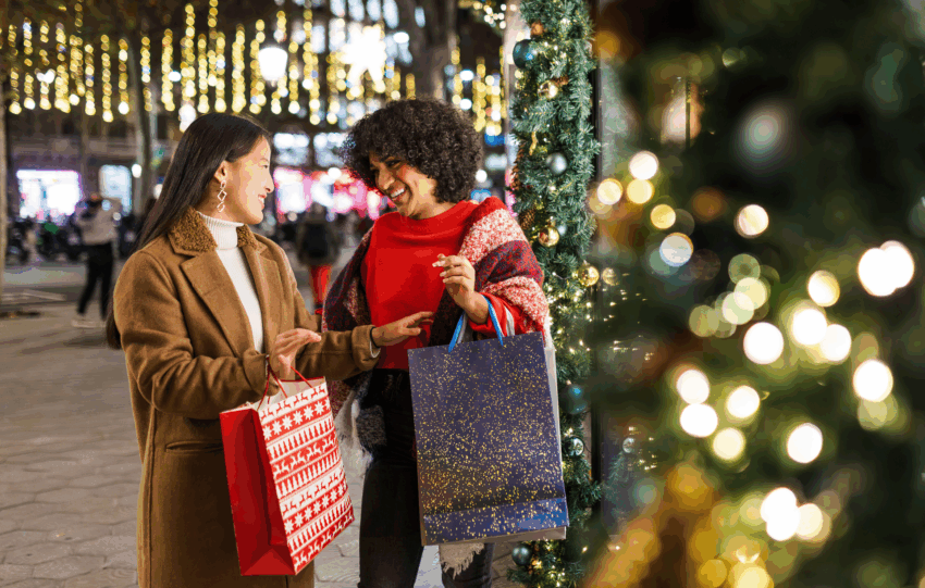 Two women holding shopping bags and smiling while standing outdoors at night surrounded by festive holiday lights.
