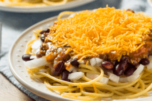 Close-up of a Cincinnati-style 5-Way chili dish on a white plate, featuring spaghetti topped with meat sauce, red kidney beans, diced white onions, and a large mound of shredded cheddar cheese.