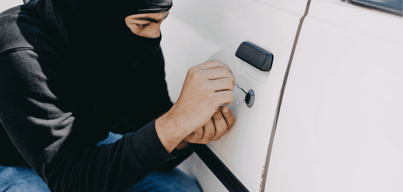 Person wearing a black balaclava attempting to pick the lock of a white car door.