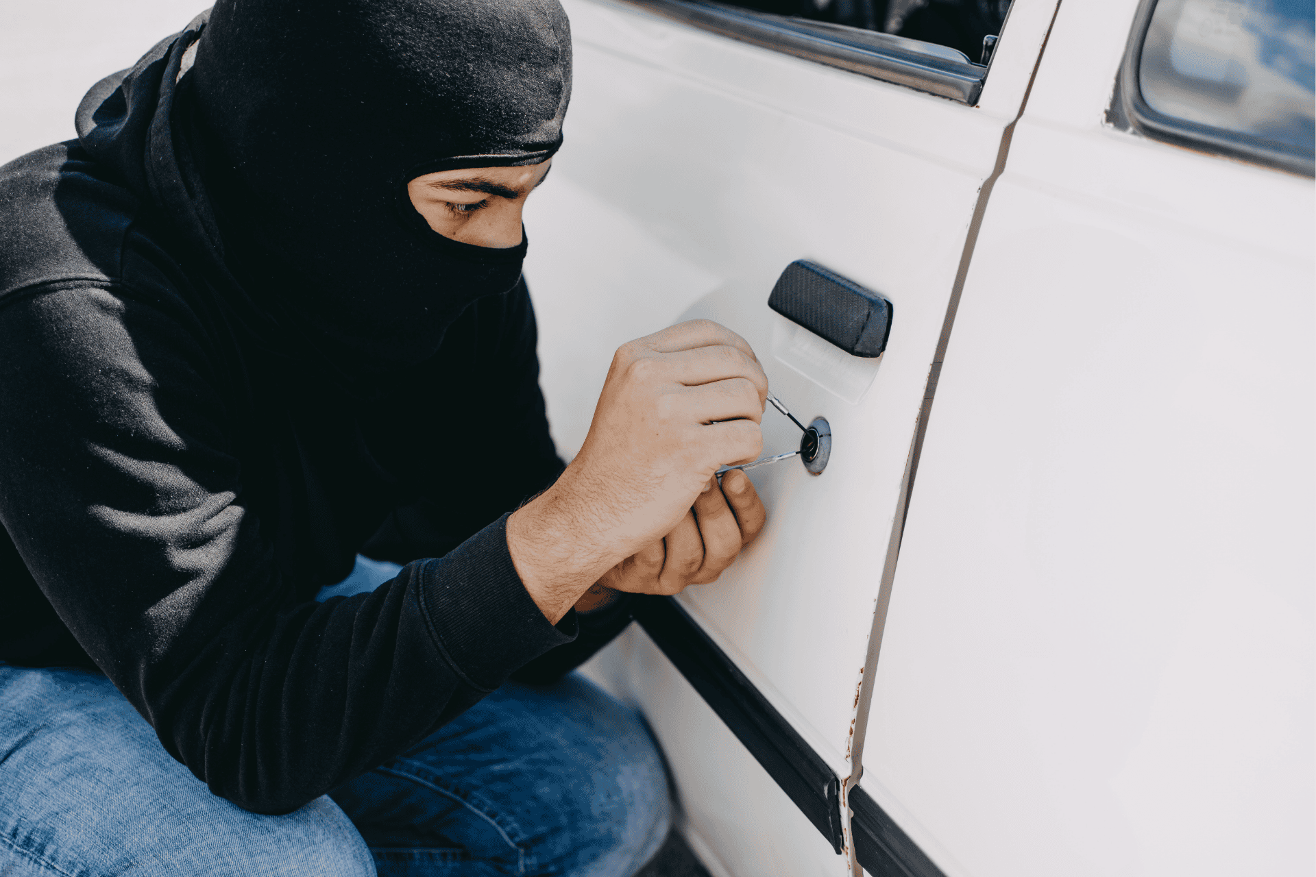 Person wearing a black balaclava attempting to pick the lock of a white car door.