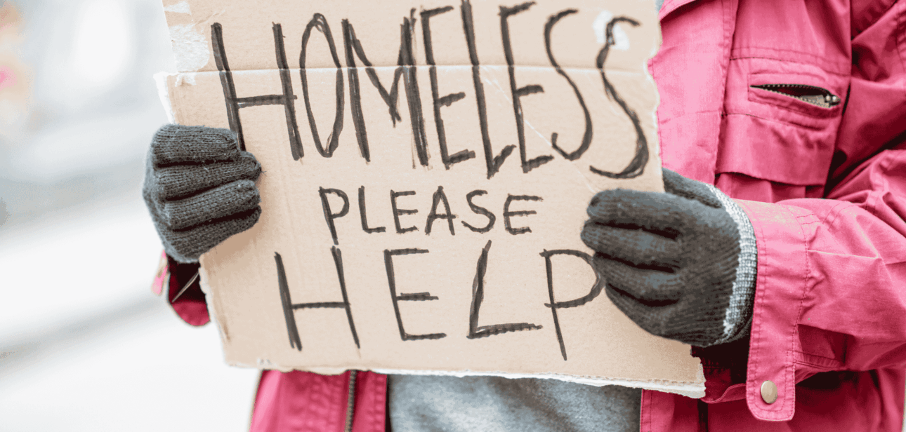 Person wearing a pink winter coat and gloves holding a handwritten cardboard sign that reads "HOMELESS PLEASE HELP."