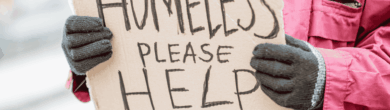 Person wearing a pink winter coat and gloves holding a handwritten cardboard sign that reads "HOMELESS PLEASE HELP."