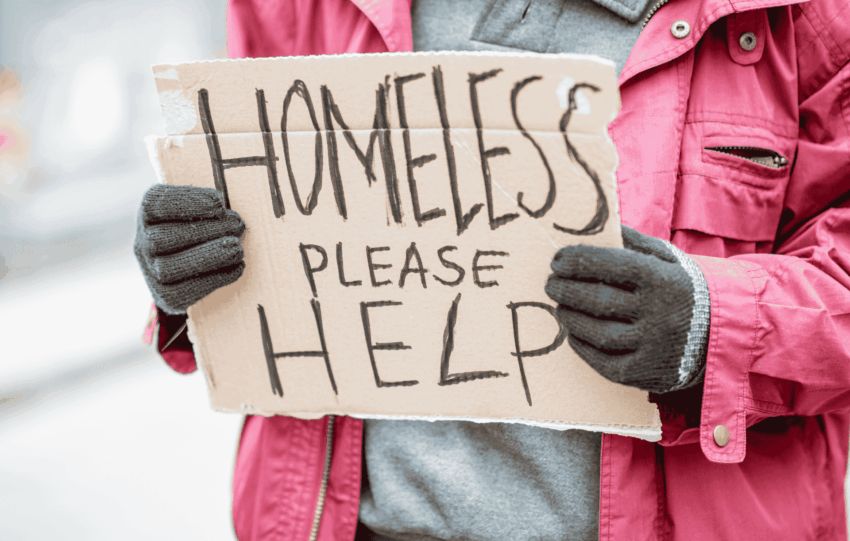 Person wearing a pink winter coat and gloves holding a handwritten cardboard sign that reads "HOMELESS PLEASE HELP."