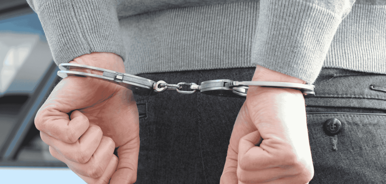 A close-up view from behind of a person’s hands secured in metal handcuffs, representing an arrest or legal custody.
