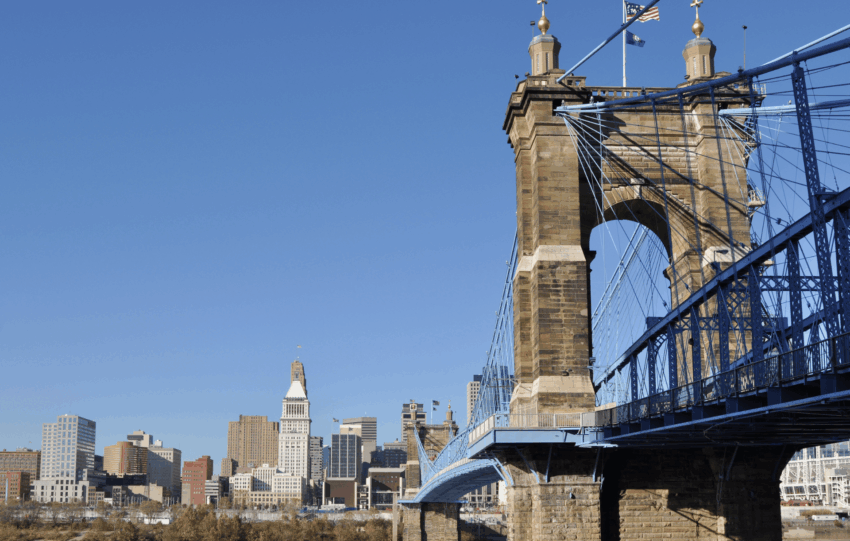 The John A. Roebling Suspension Bridge spanning the Ohio River with the Cincinnati skyline in the background under a blue sky.