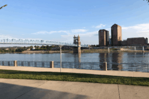 The historic blue John A. Roebling Suspension Bridge spanning the Ohio River, viewed from a paved walking path on the Cincinnati riverfront.
