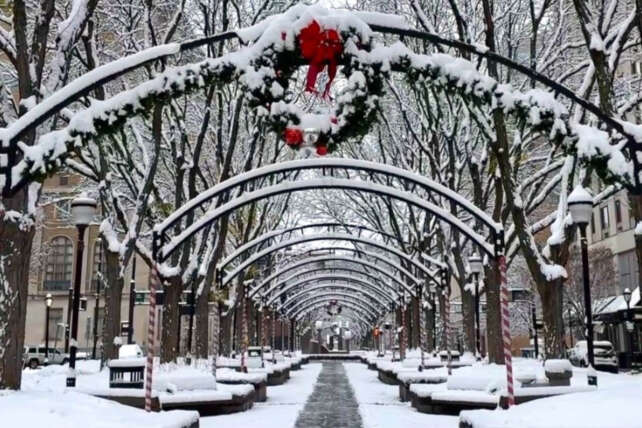 Snow covered pedestrian walkway in Cincinnati featuring arched trellises with holiday wreaths and snow dusted benches.