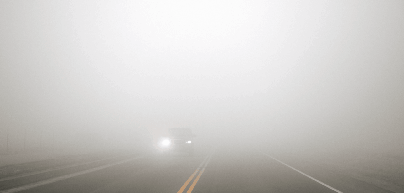 Dense fog obscuring a two-lane road with oncoming car headlights, illustrating the Cincinnati weather forecast for New Year's.