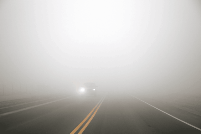 Dense fog obscuring a two-lane road with oncoming car headlights, illustrating the Cincinnati weather forecast for New Year's.