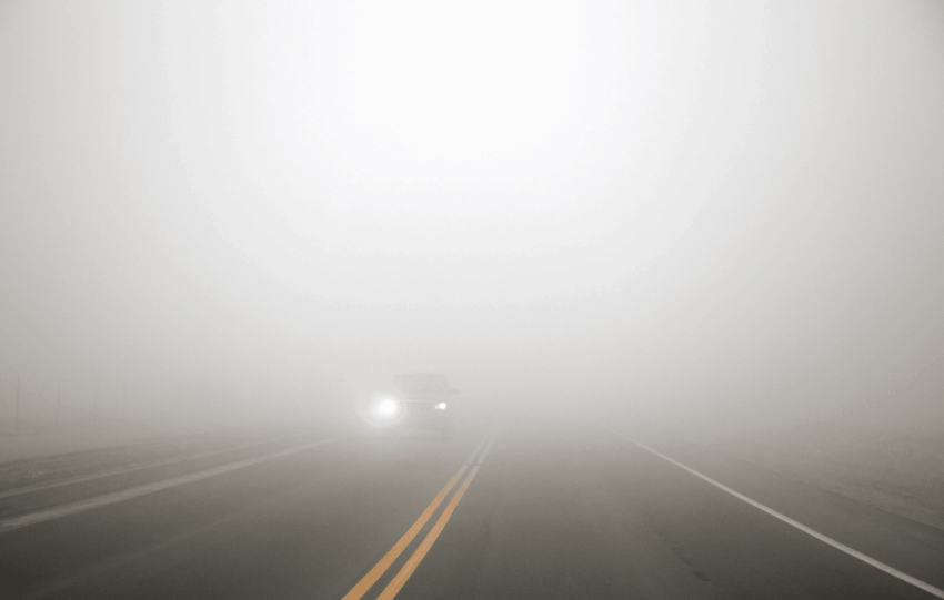 Dense fog obscuring a two-lane road with oncoming car headlights, illustrating the Cincinnati weather forecast for New Year's.