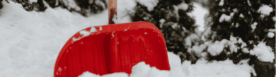 Red snow shovel stuck in a pile of deep, fresh snow with snow-covered evergreen trees in the background.