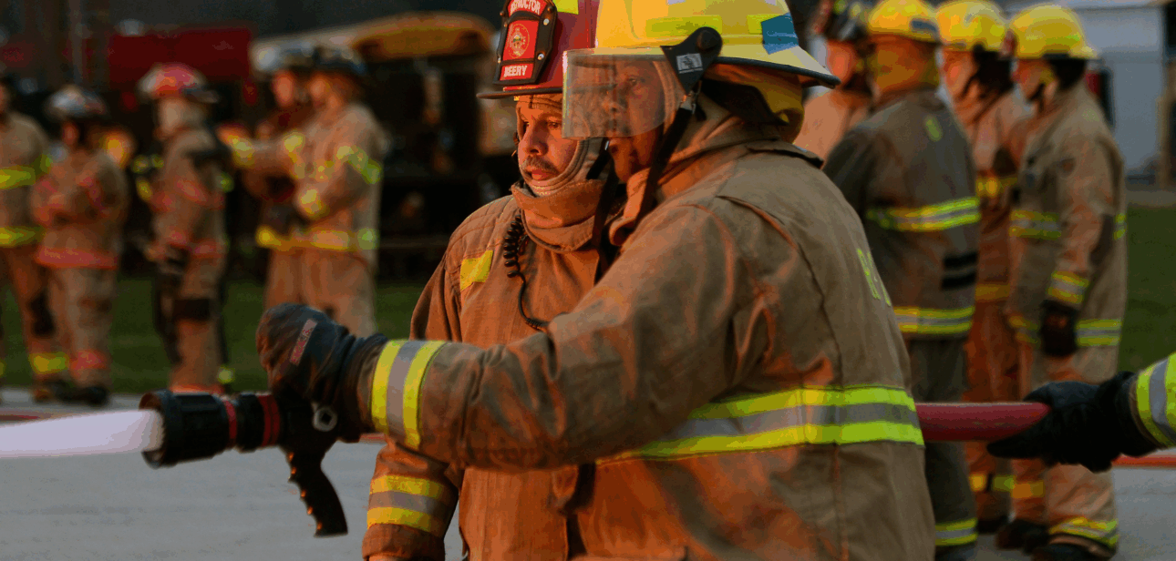 A firefighter in tan turnout gear and a yellow helmet holding a hose, with a group of other firefighters standing in the background.