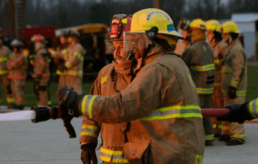 A firefighter in tan turnout gear and a yellow helmet holding a hose, with a group of other firefighters standing in the background.