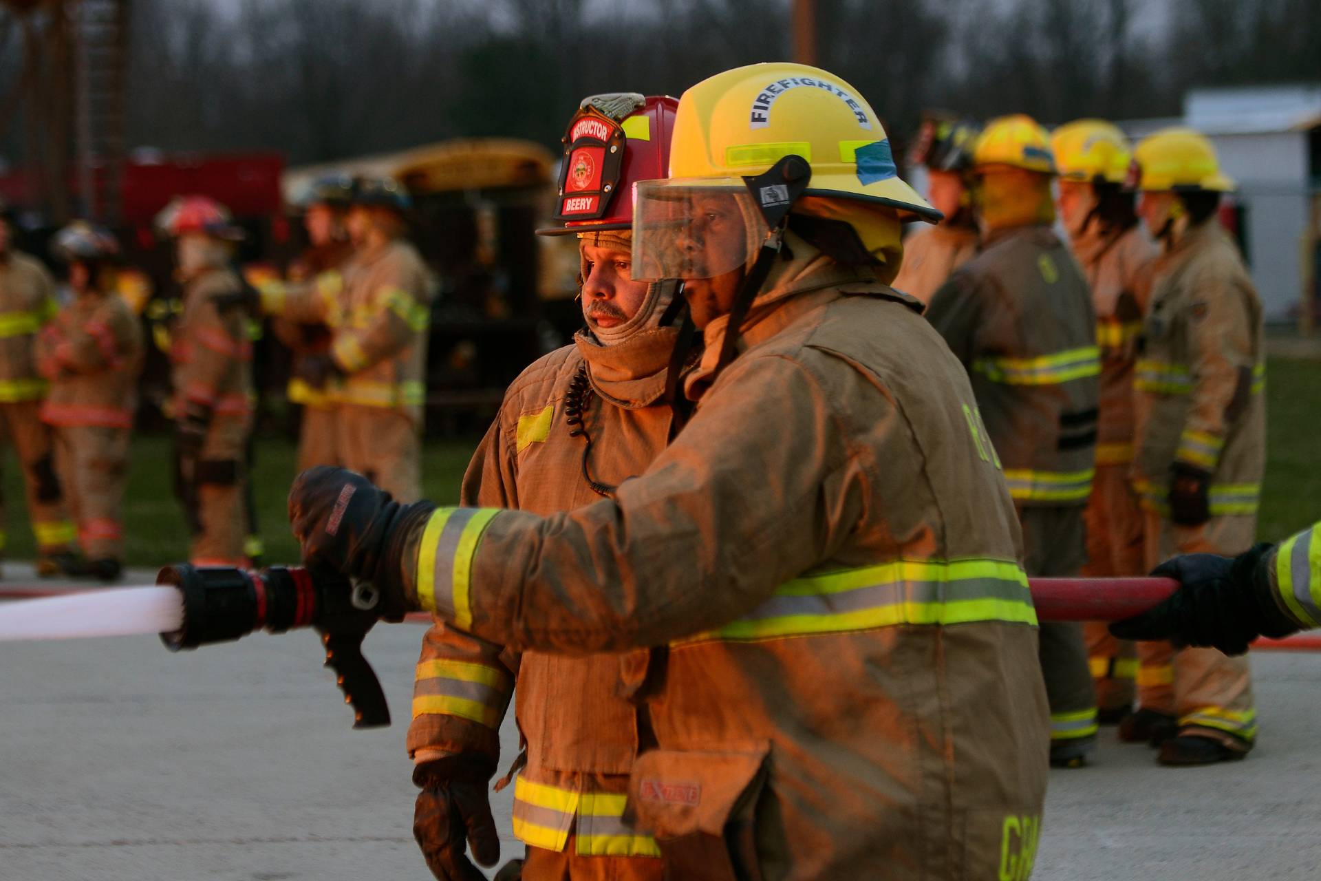 A firefighter in tan turnout gear and a yellow helmet holding a hose, with a group of other firefighters standing in the background.