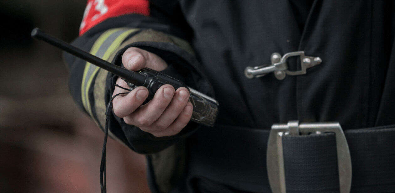 Close-up of a firefighter in uniform holding a handheld two-way radio device against a blurred background.
