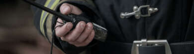 Close-up of a firefighter in uniform holding a handheld two-way radio device against a blurred background.