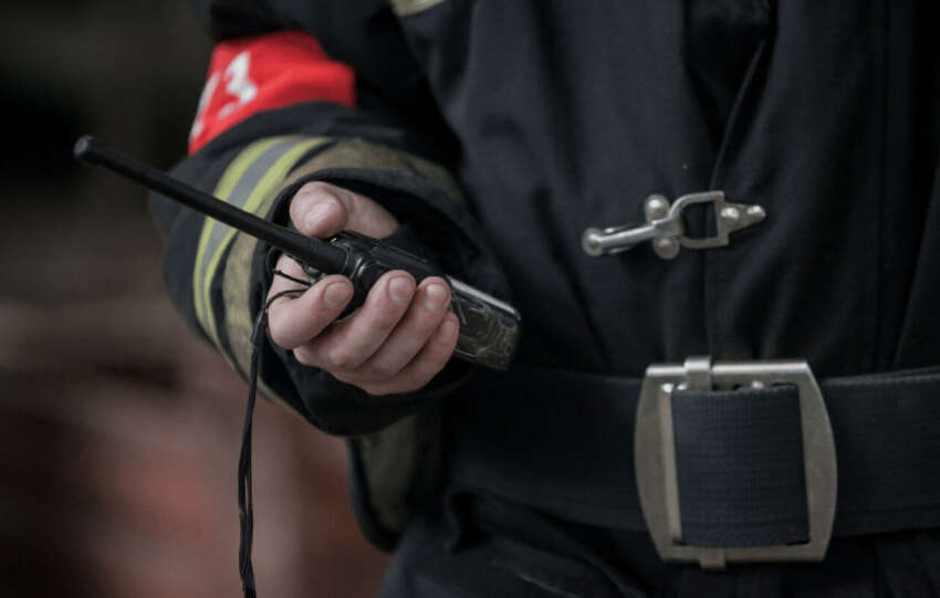 Close-up of a firefighter in uniform holding a handheld two-way radio device against a blurred background.
