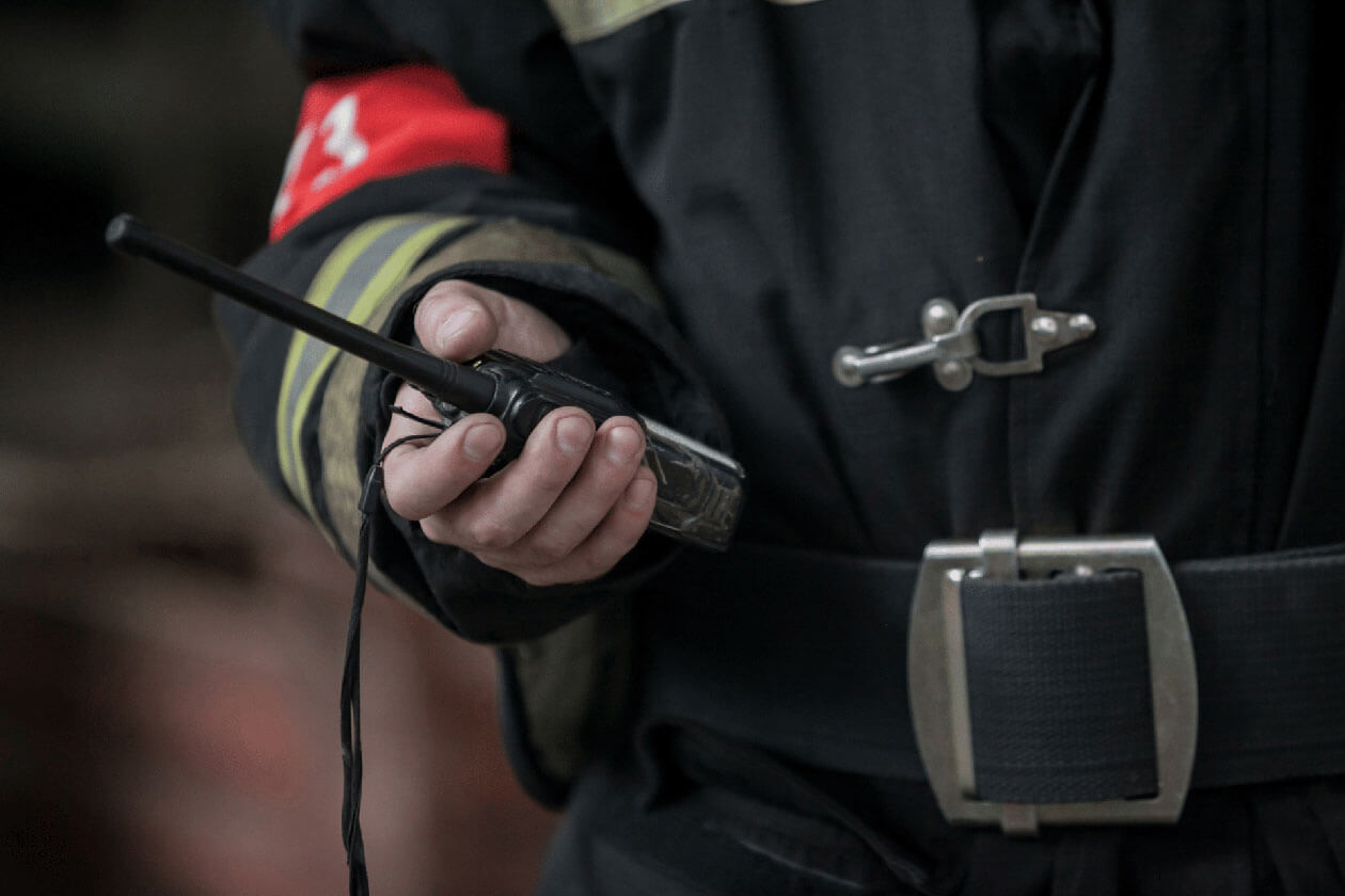 Close-up of a firefighter in uniform holding a handheld two-way radio device against a blurred background.