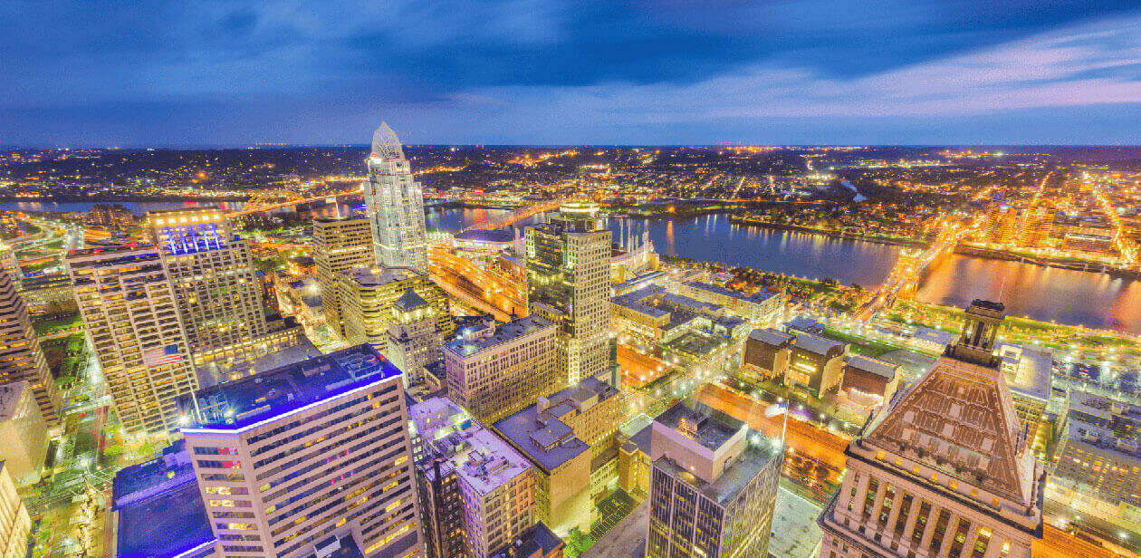 High-angle twilight view of the Cincinnati skyline with illuminated skyscrapers, the Great American Tower, and the Ohio River in the background.