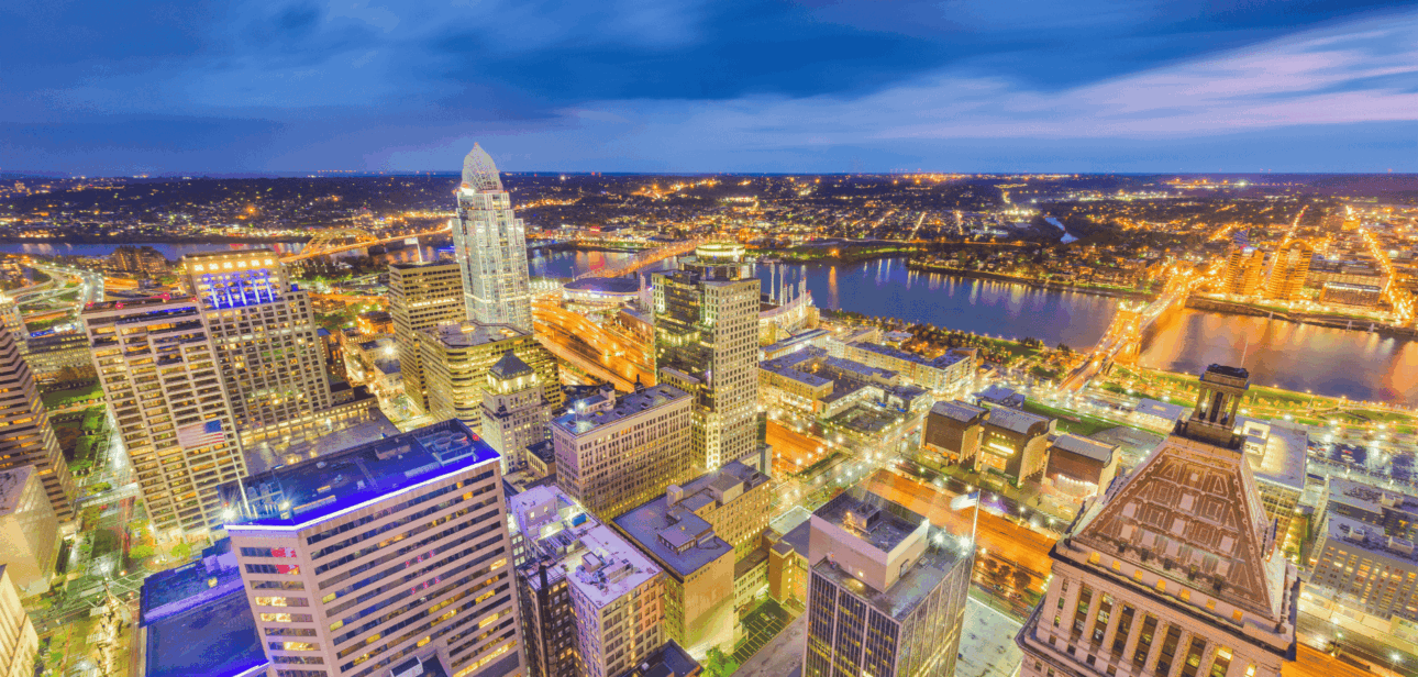 High-angle twilight view of the Cincinnati skyline with illuminated skyscrapers, the Great American Tower, and the Ohio River in the background.