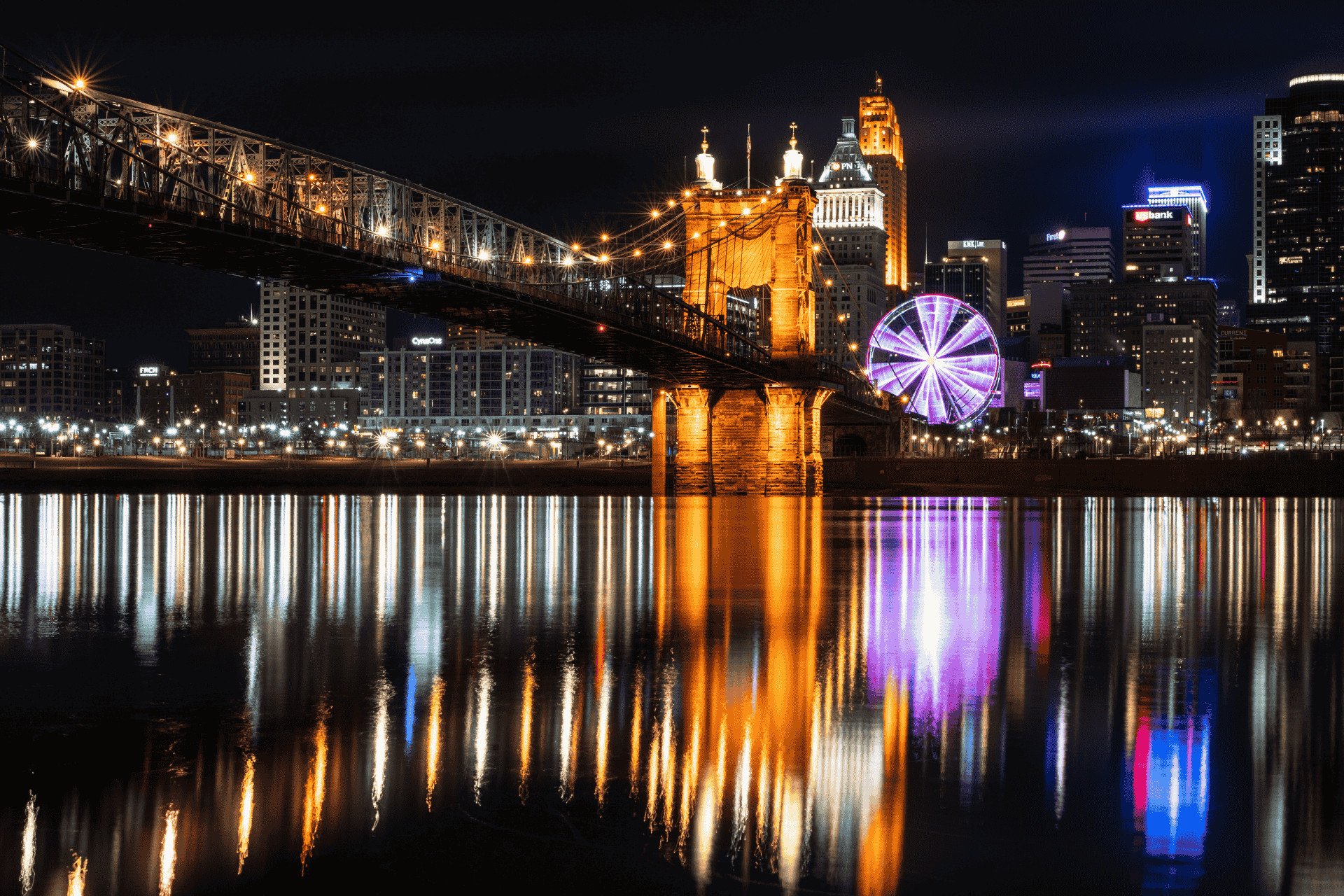 Nighttime view of the Cincinnati skyline and illuminated Roebling Suspension Bridge reflecting on the Ohio River.
