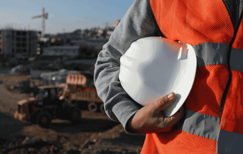 Construction worker in an orange safety vest holding a white hard hat in front of a blurred construction site with a crane, representing thriving industries in Cincinnati.