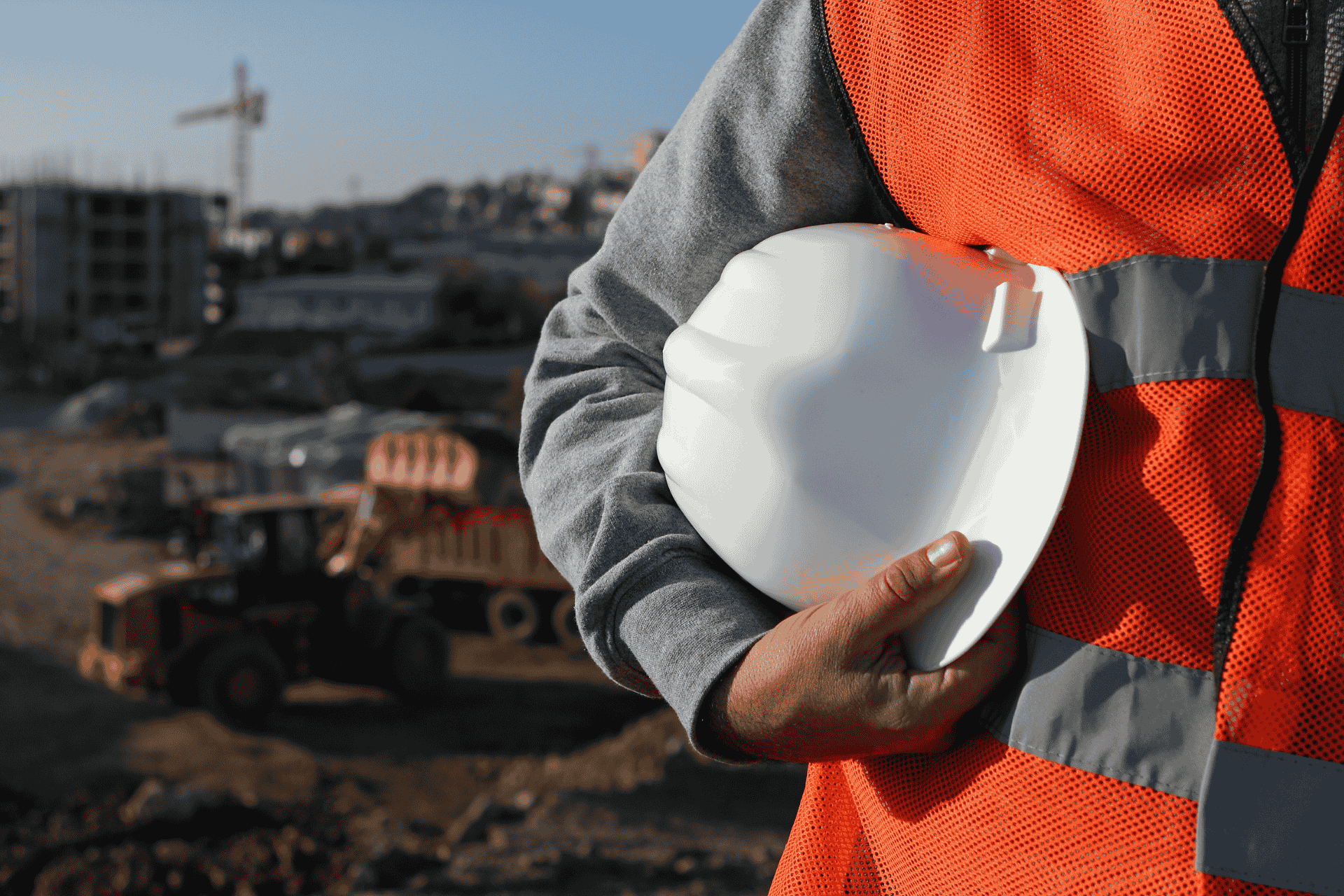 Construction worker in an orange safety vest holding a white hard hat in front of a blurred construction site with a crane, representing thriving industries in Cincinnati.