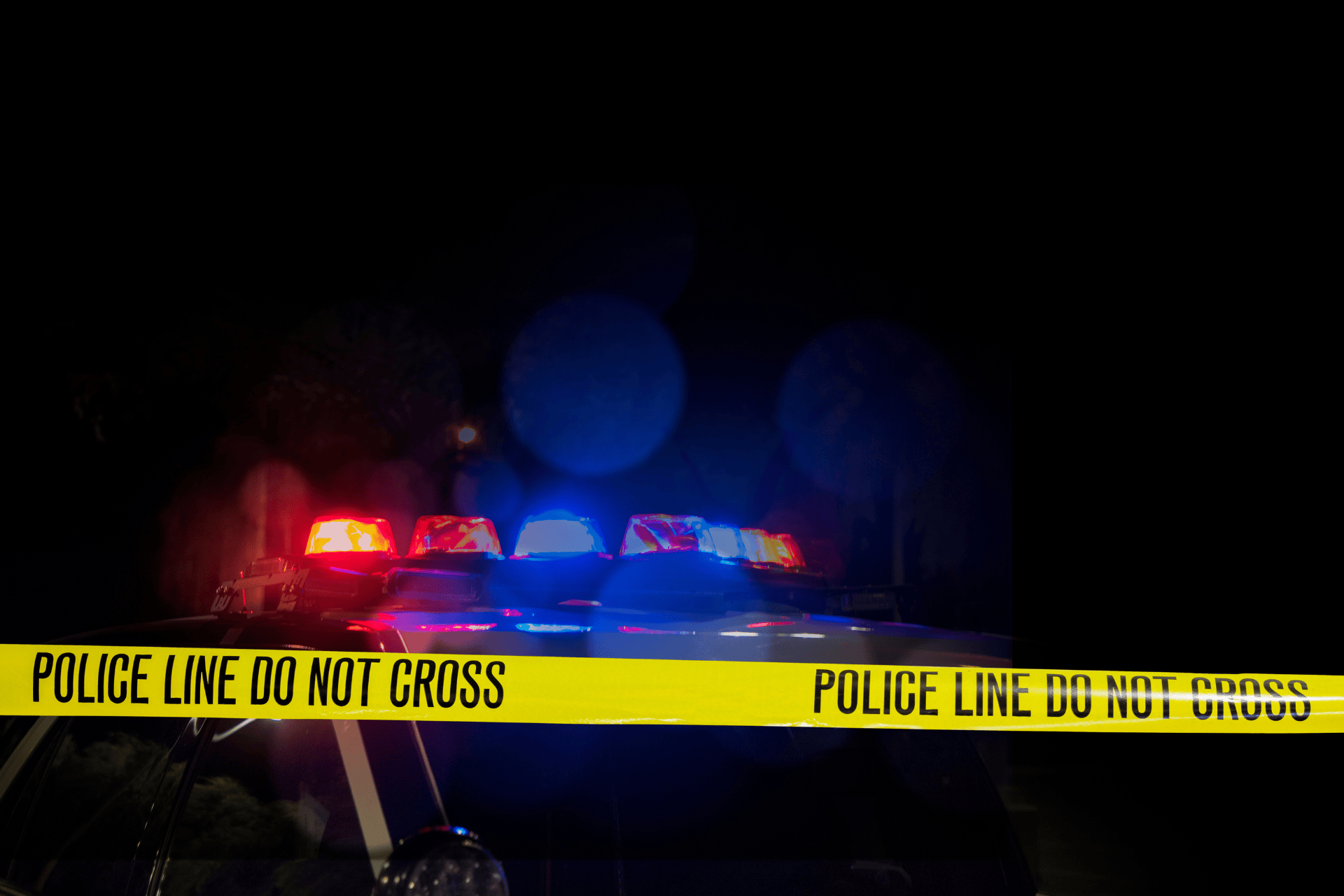 Police cruiser lights flashing blue and red behind yellow do not cross tape at night, illustrating a shooting investigation in Cincinnati's West End.