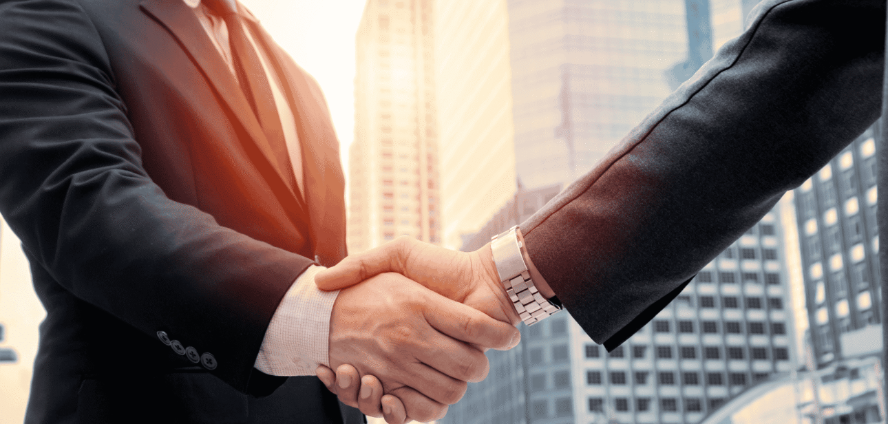 Two business professionals in suits shaking hands against a sunset city skyline, representing a corporate real estate acquisition.