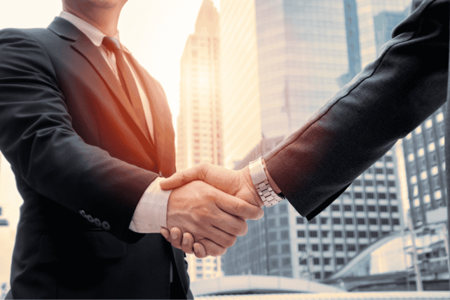 Two business professionals in suits shaking hands against a sunset city skyline, representing a corporate real estate acquisition.