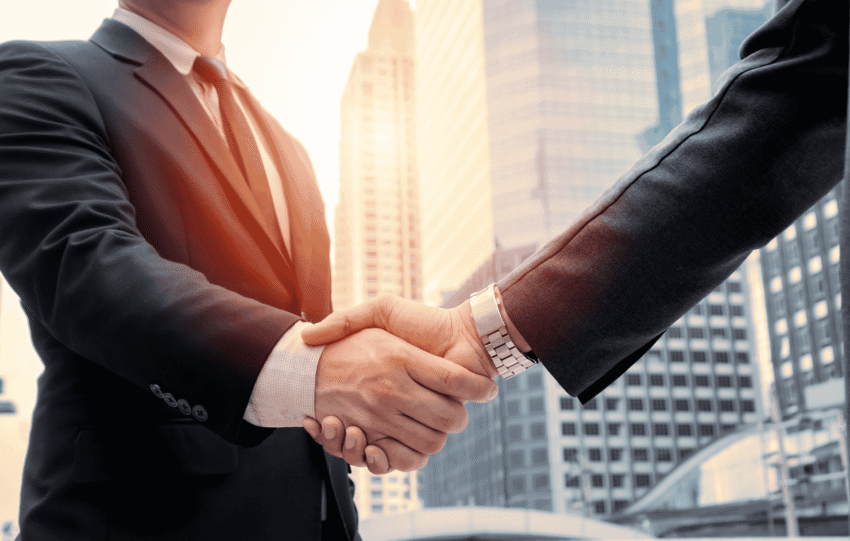 Two business professionals in suits shaking hands against a sunset city skyline, representing a corporate real estate acquisition.