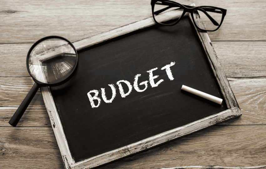 A magnifying glass and eyeglasses resting on a chalkboard with the word "BUDGET" written in chalk.