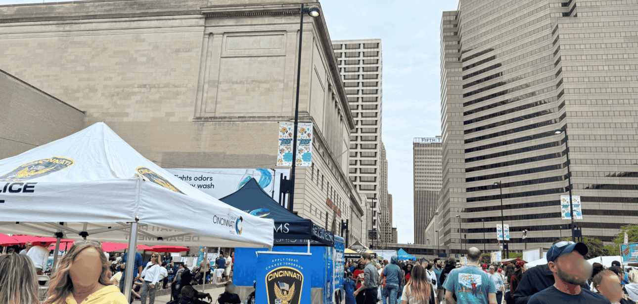 Large crowd in the City of Cincinnati during the Taste of Cincinnati 2025 featuring local police booths and downtown skyscrapers.
