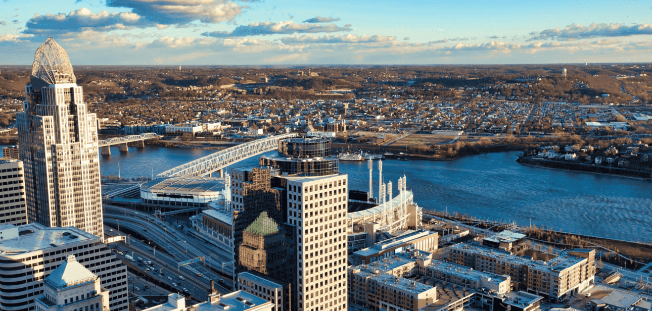 An aerial panoramic view of the Cincinnati skyline at sunset, featuring prominent skyscrapers and the Ohio River riverfront under a clear blue sky.