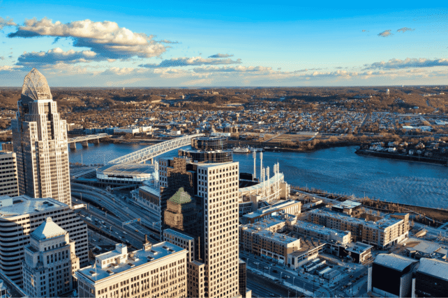 An aerial panoramic view of the Cincinnati skyline at sunset, featuring prominent skyscrapers and the Ohio River riverfront under a clear blue sky.