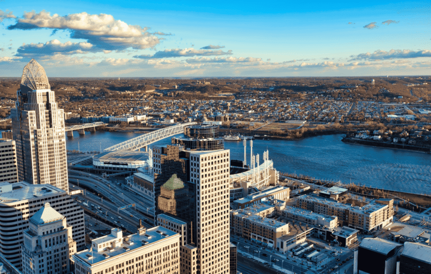 An aerial panoramic view of the Cincinnati skyline at sunset, featuring prominent skyscrapers and the Ohio River riverfront under a clear blue sky.