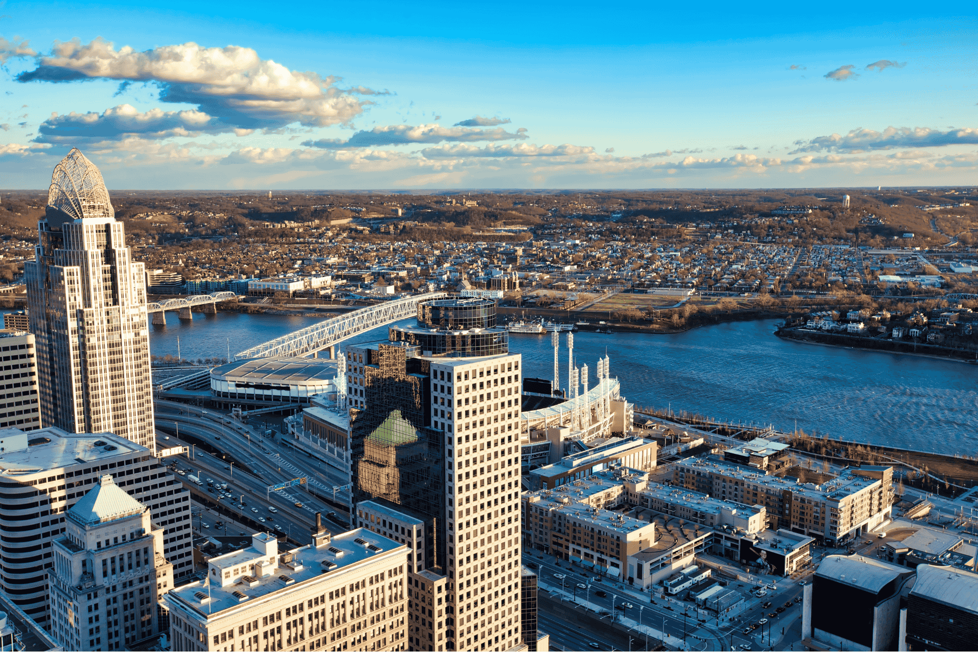 An aerial panoramic view of the Cincinnati skyline at sunset, featuring prominent skyscrapers and the Ohio River riverfront under a clear blue sky.