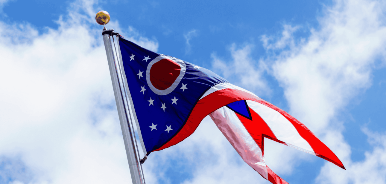Ohio state flag waving on a flagpole against a blue sky with white clouds.