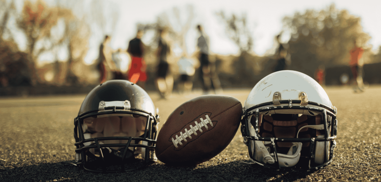 Two football helmets and a football resting on a grassy field with blurred players practicing in the background during a team workout.
